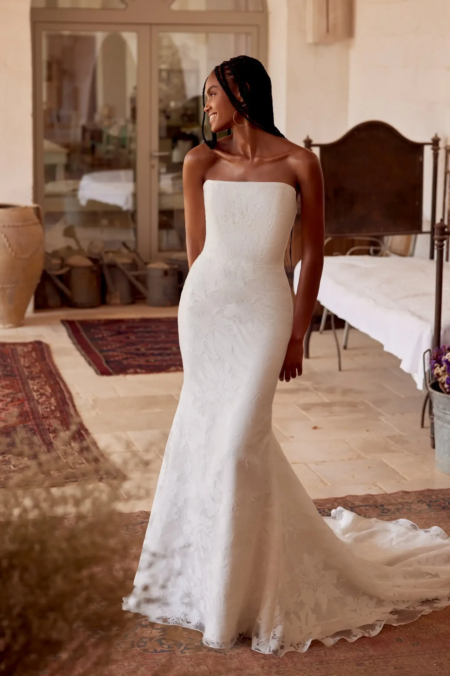 A woman in a strapless, fitted white lace wedding gown stands smiling on a vintage rug in a rustic setting. Soft, natural light creates an elegant and serene feel.