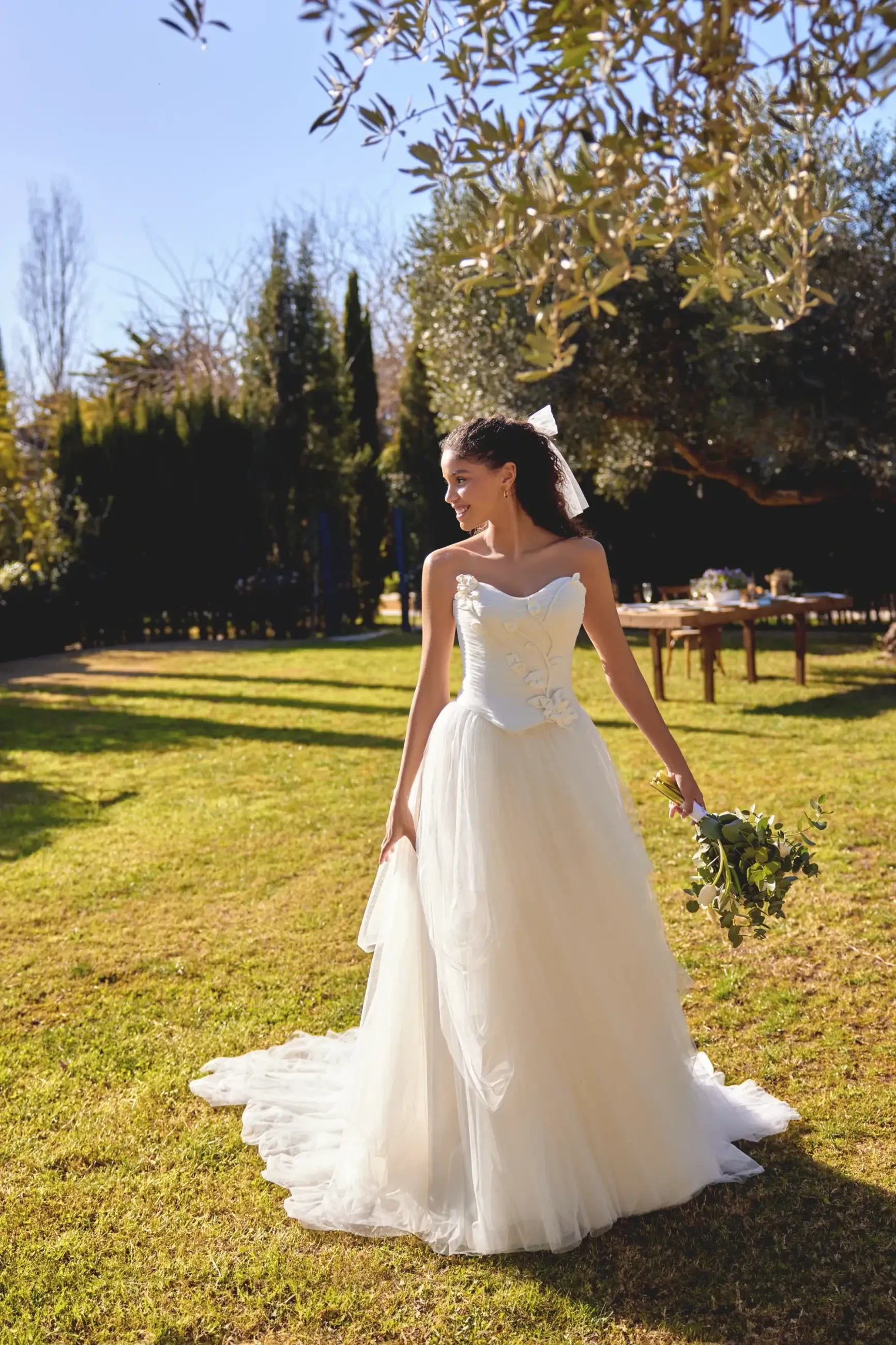 A bride in a flowing white gown stands on a sunlit lawn, holding a bouquet. She smiles to the side, with trees and a banquet table in the background.