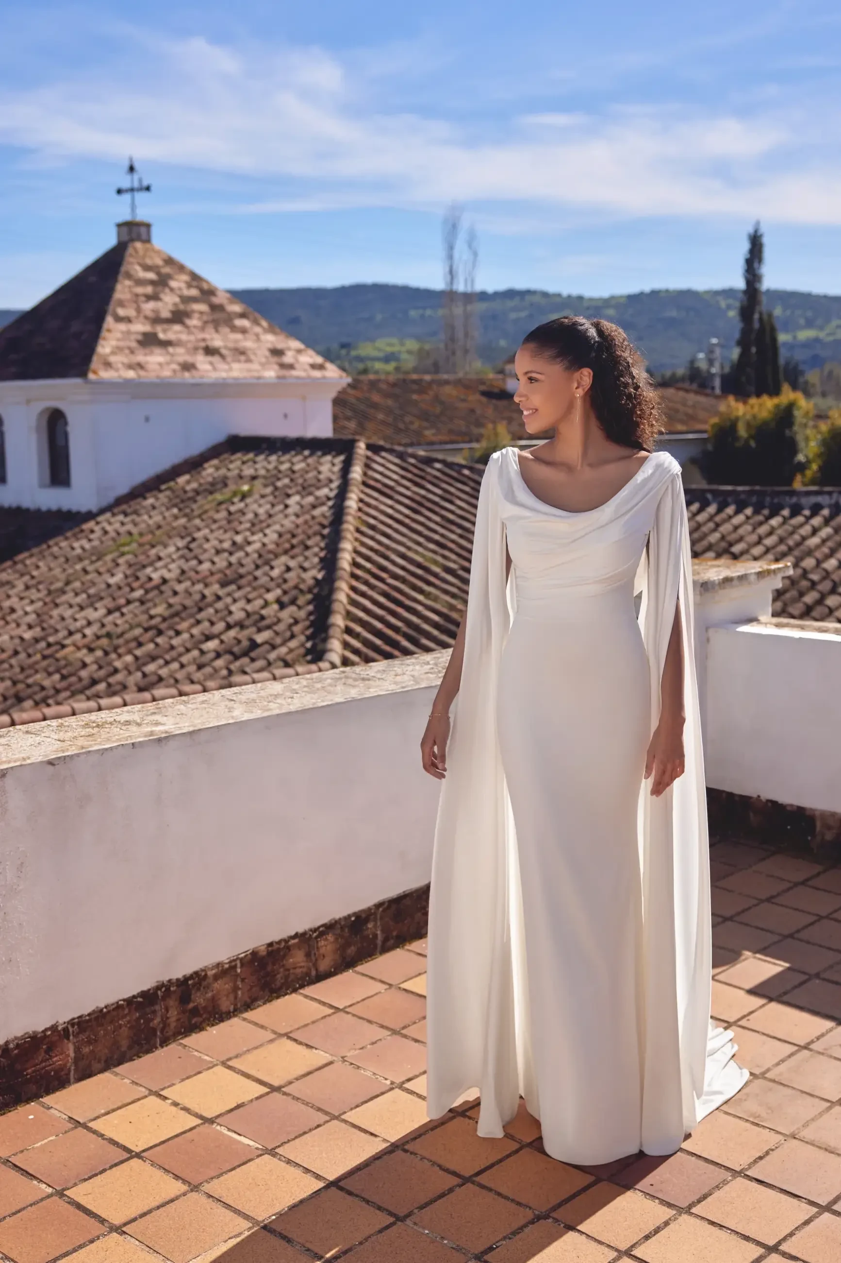 A woman wearing a long, white dress with a cape stands on a terrace with a scenic view of rooftops and hills in the background.