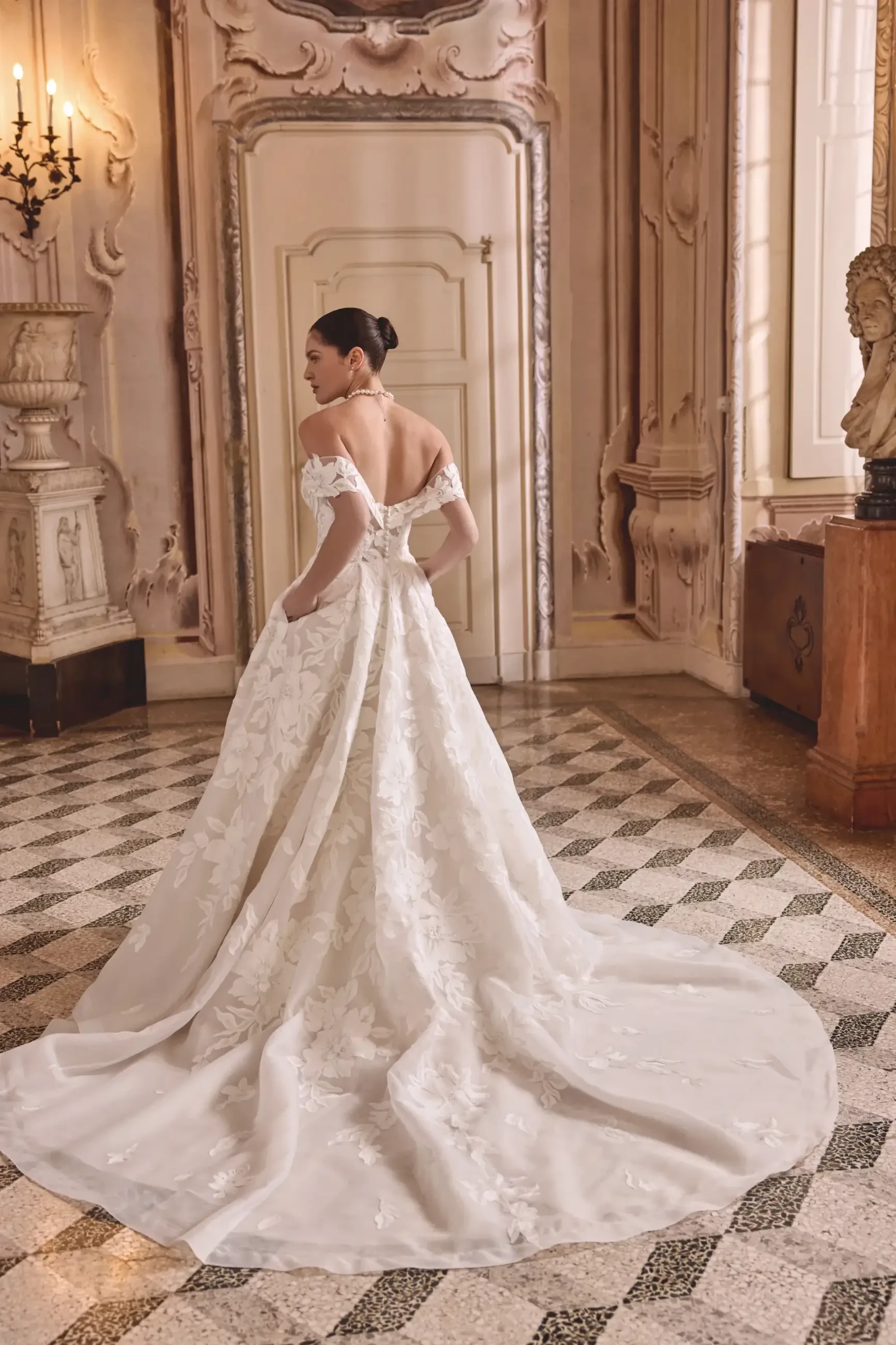 A bride in an elegant off-shoulder gown with floral patterns stands in an ornate room, evoking a timeless, romantic ambiance. The long train flows over a checkered floor.