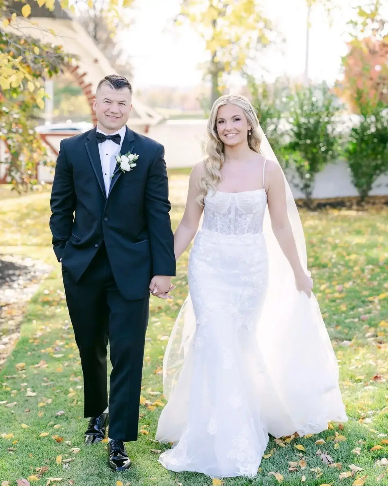 A bride and groom walking hand in hand outdoors, with trees and greenery in the background, dressed in formal wedding attire.