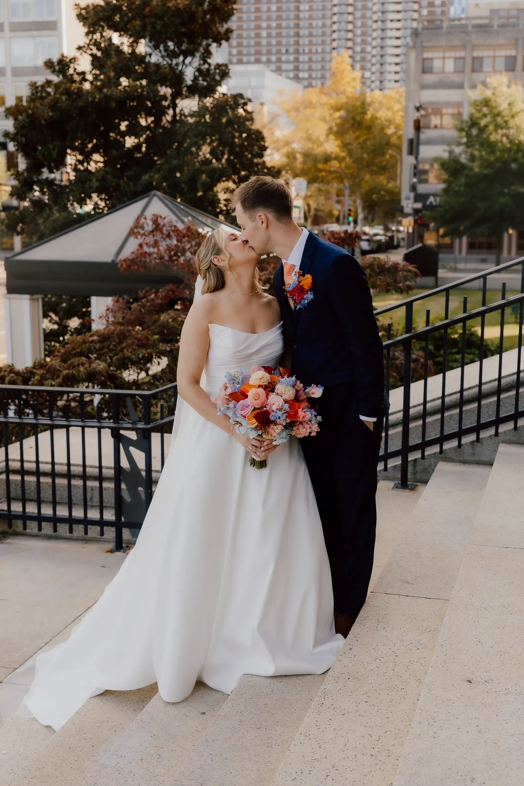 A couple in wedding attire kisses outdoors, holding a bouquet of flowers and a leash attached to a black dog. The background features a red door and brick walkway.