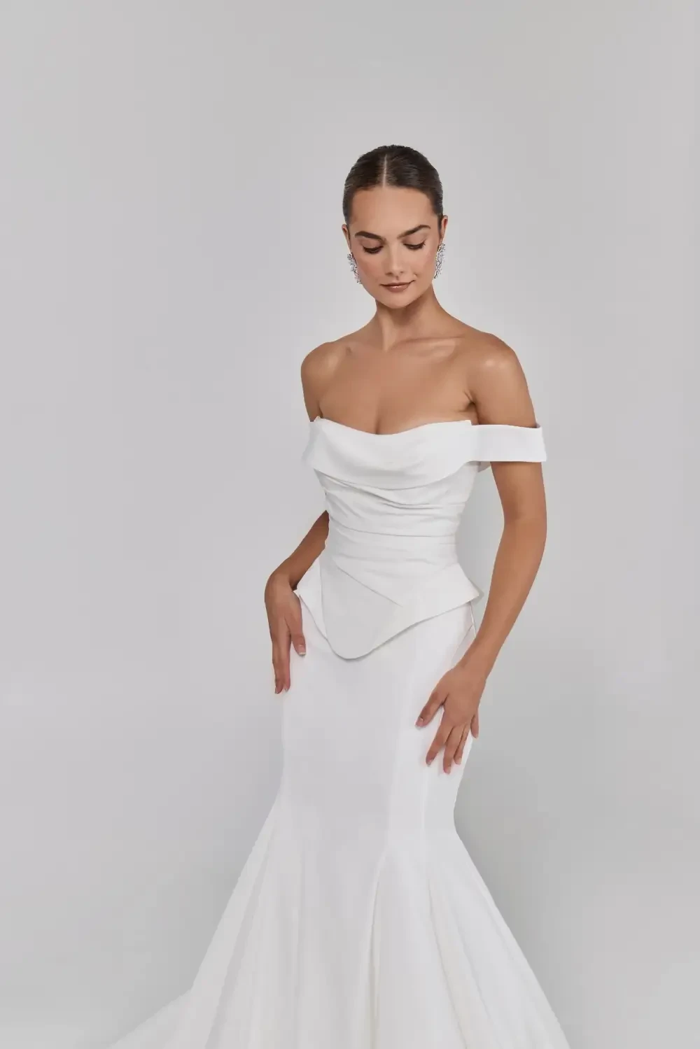 A woman in a fitted off-the-shoulder white dress, standing gracefully with her hands at her waist.