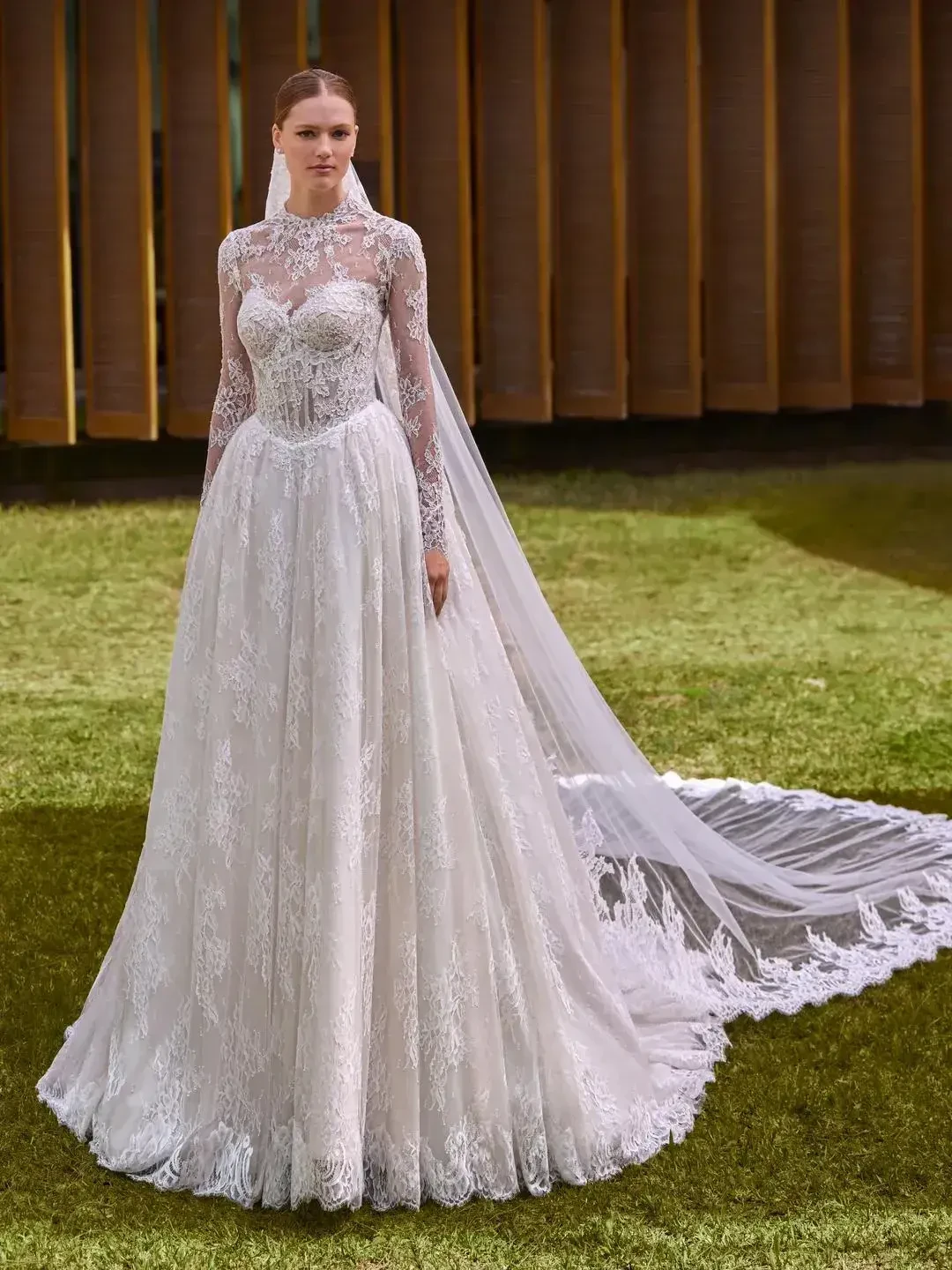 A bride wearing a long-sleeved lace wedding gown with a full skirt and train, standing on green grass.