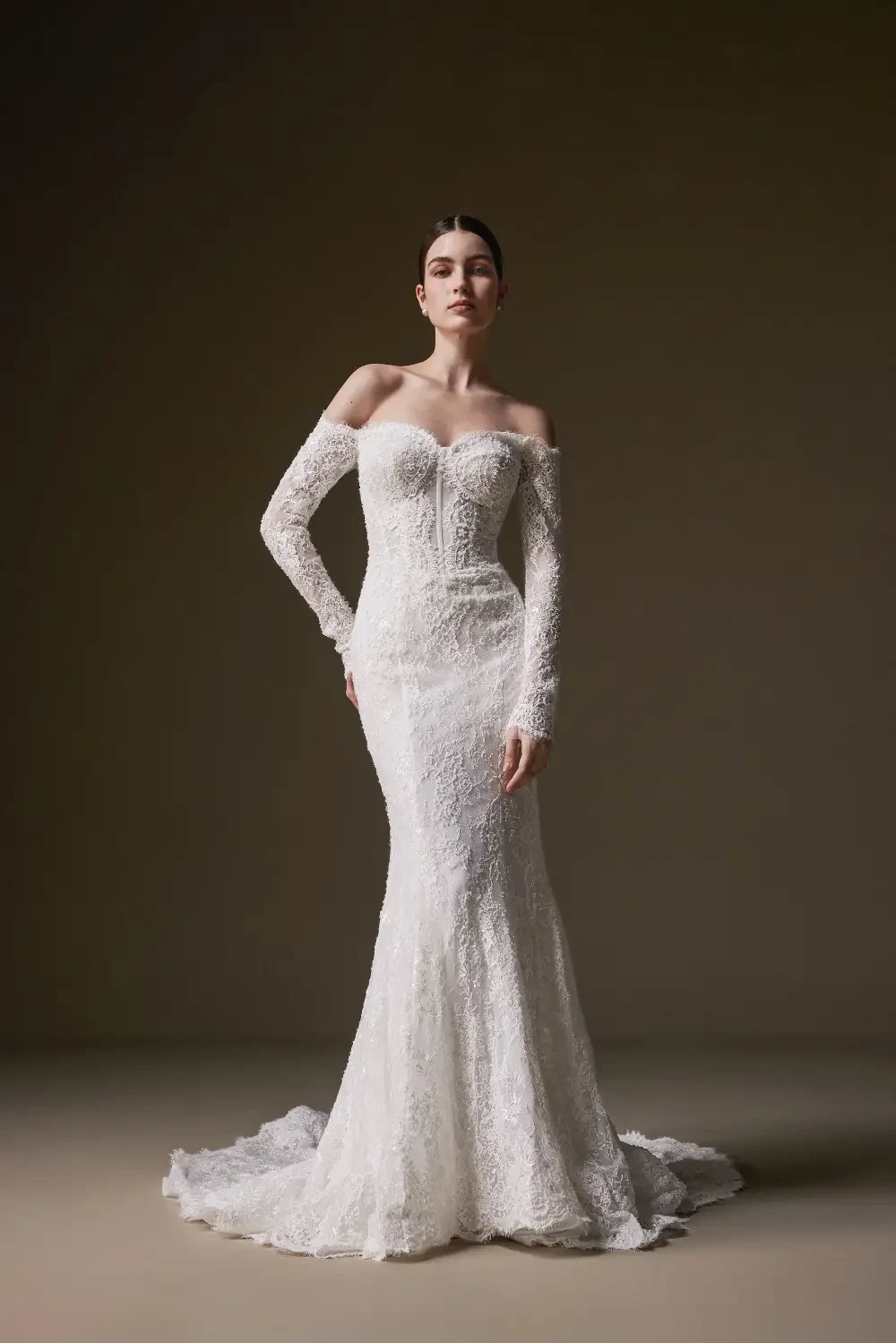 A model wearing an elegant off-the-shoulder lace wedding dress with long sleeves, standing against a dark background.