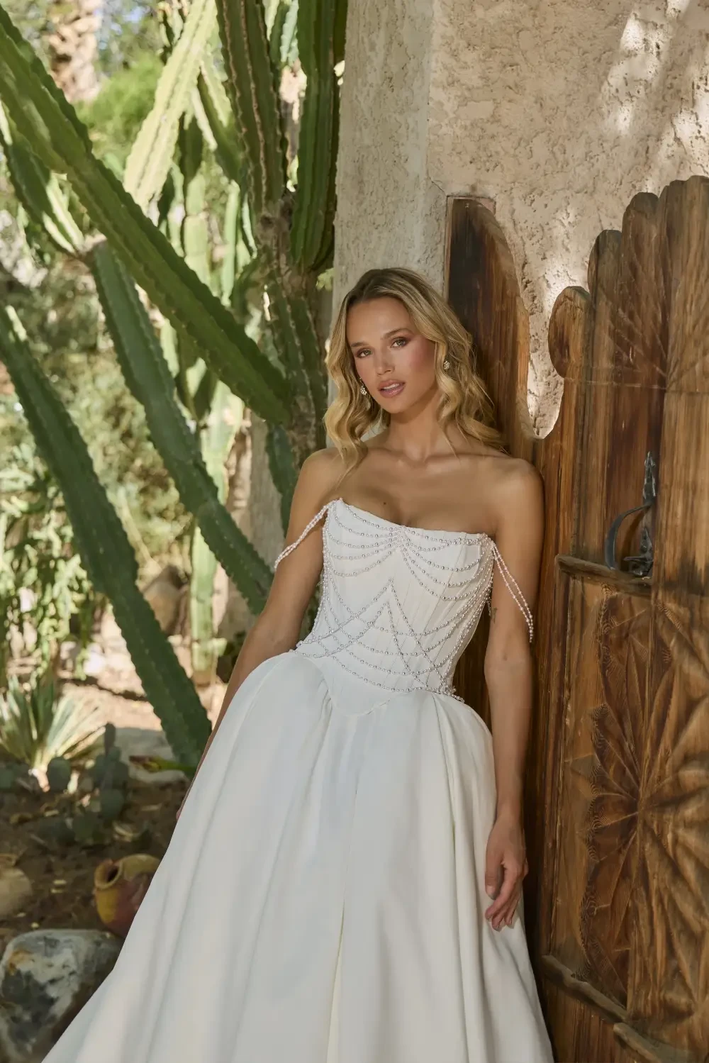 A woman in a strapless white gown stands beside a rustic wooden door, surrounded by cacti and greenery.