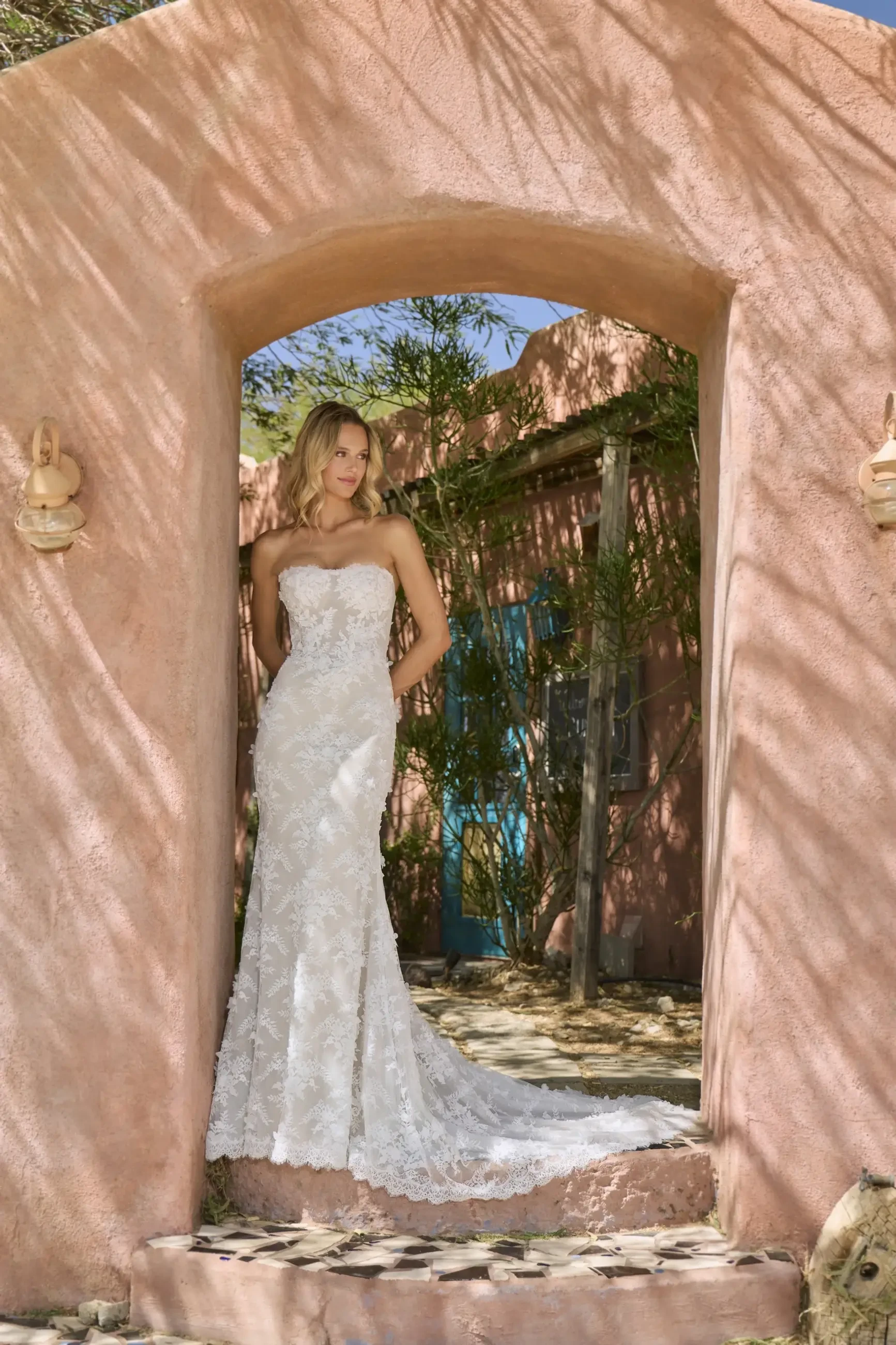 A bride wearing a strapless lace wedding dress stands under an archway with pink walls, surrounded by greenery.