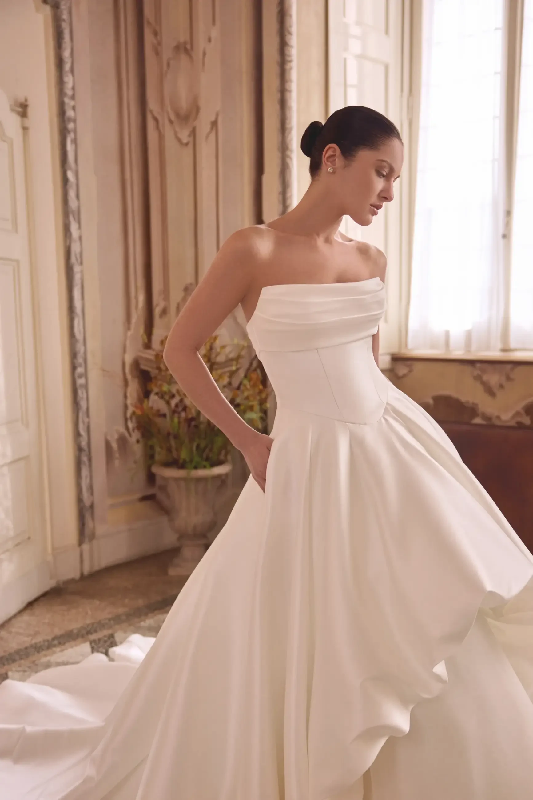 A woman in an elegant, strapless white wedding gown stands in a sunlit room with ornate decor. She conveys a serene, contemplative mood.