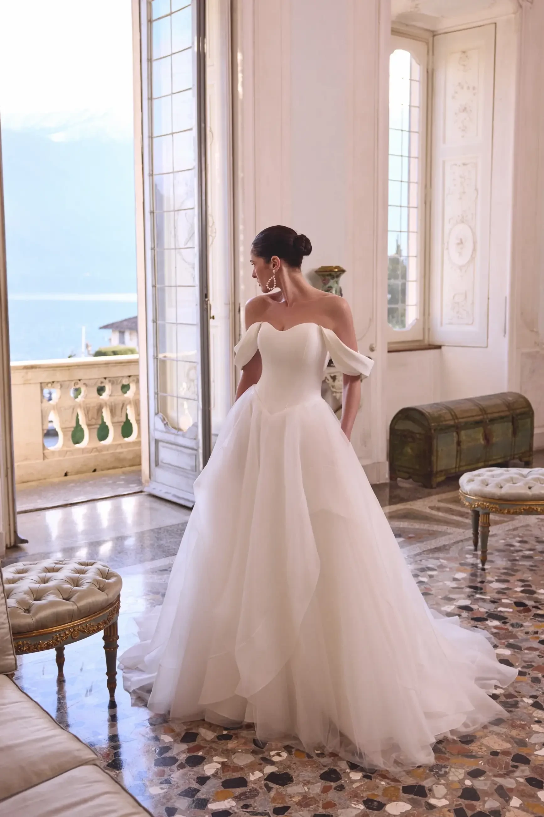 A woman in a wedding dress stands elegantly in a luxurious room with large windows and intricate flooring, looking out towards a scenic view.