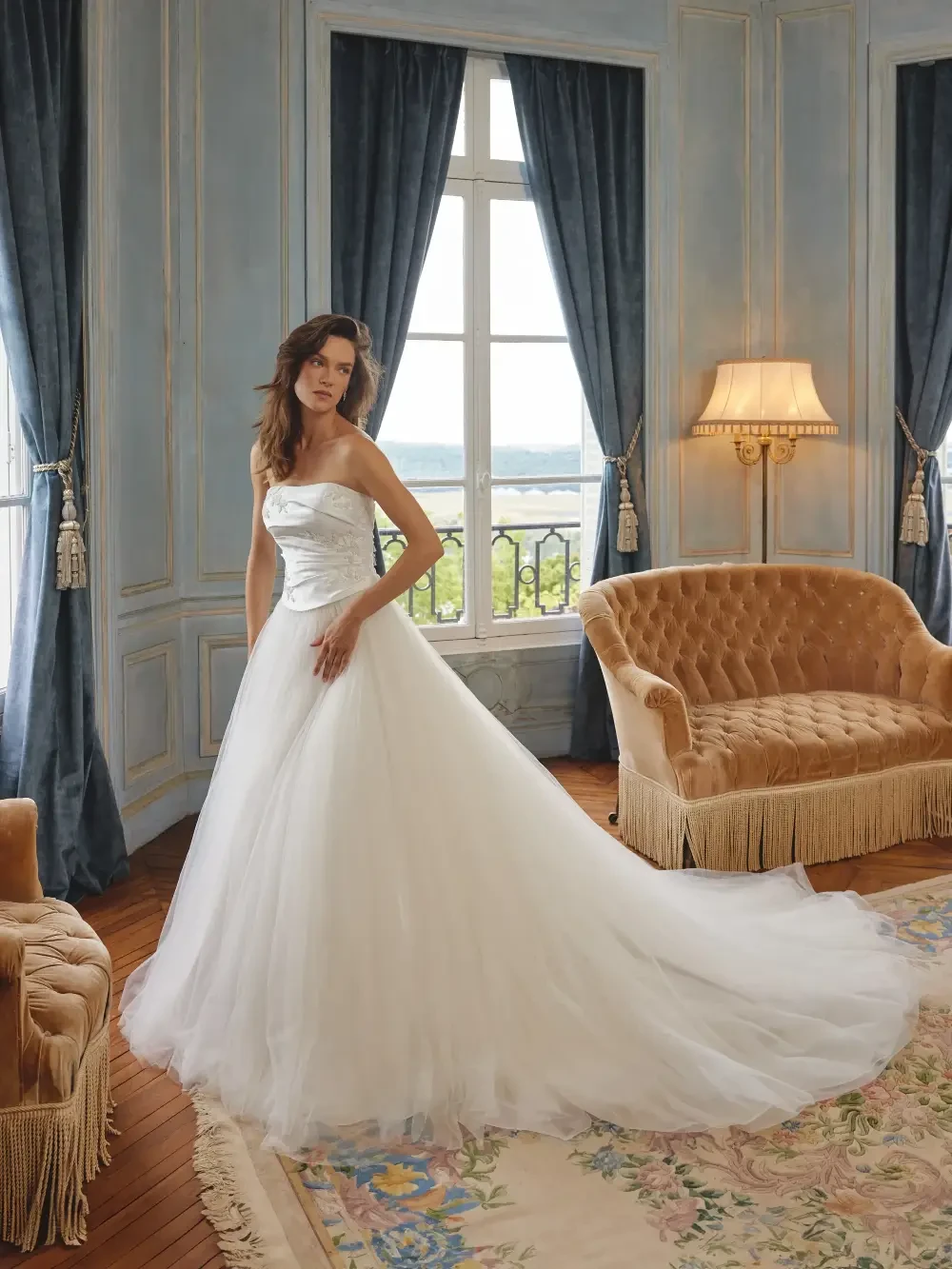 A bride in a white wedding gown stands elegantly in a beautifully decorated room with blue curtains and a vintage-style sofa.