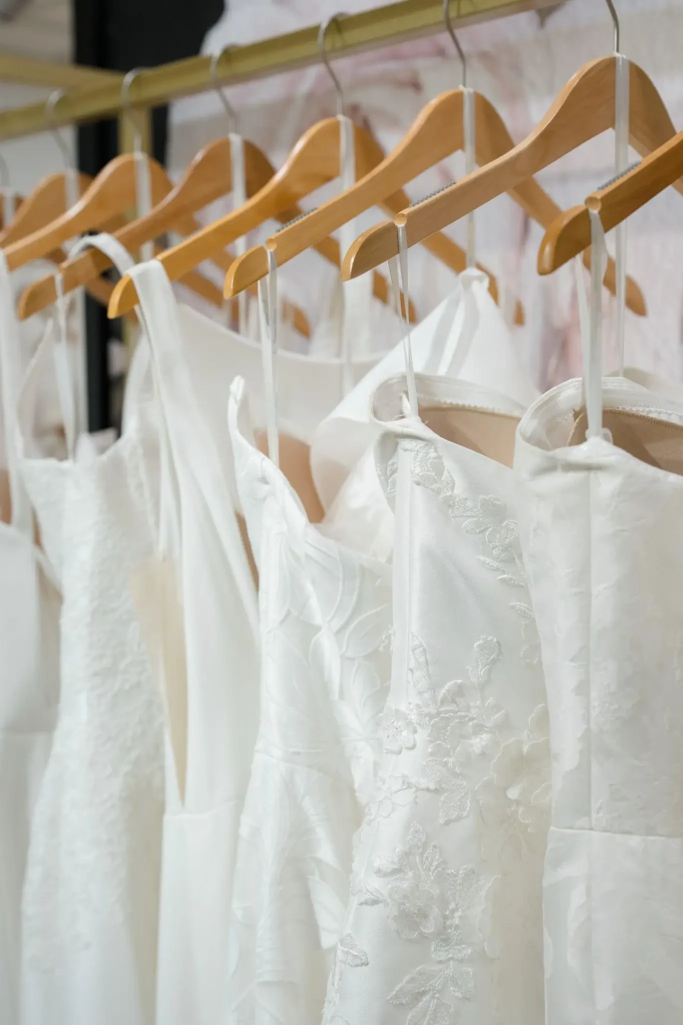A close-up of white and ivory wedding gowns with lace and floral detailing hanging on wooden hangers inside a bridal boutique.