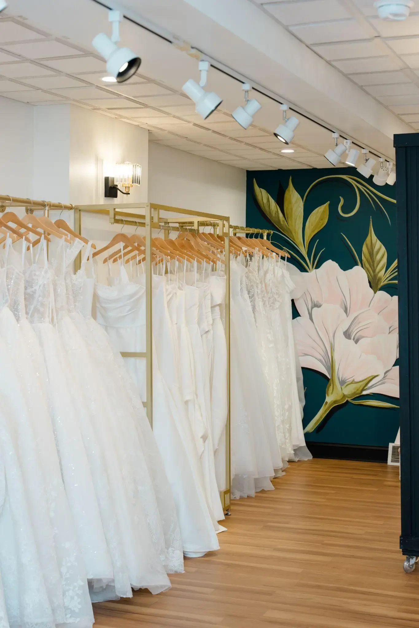 Rows of white and ivory wedding gowns displayed on gold racks inside the Le Bella Donna bridal boutique, with a floral mural in the background.
