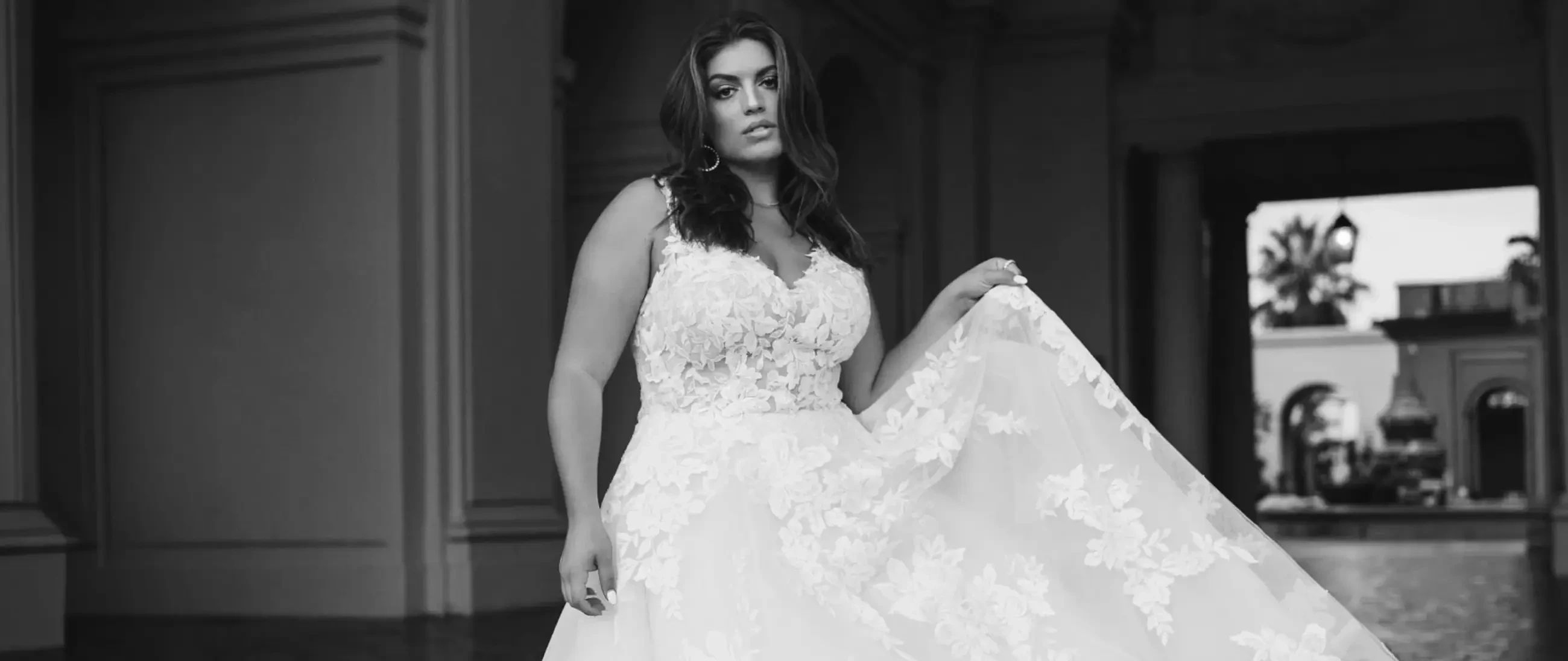 Model wearing a plus-size lace wedding gown with a V-neckline, floral appliqués, and a full flowing skirt.