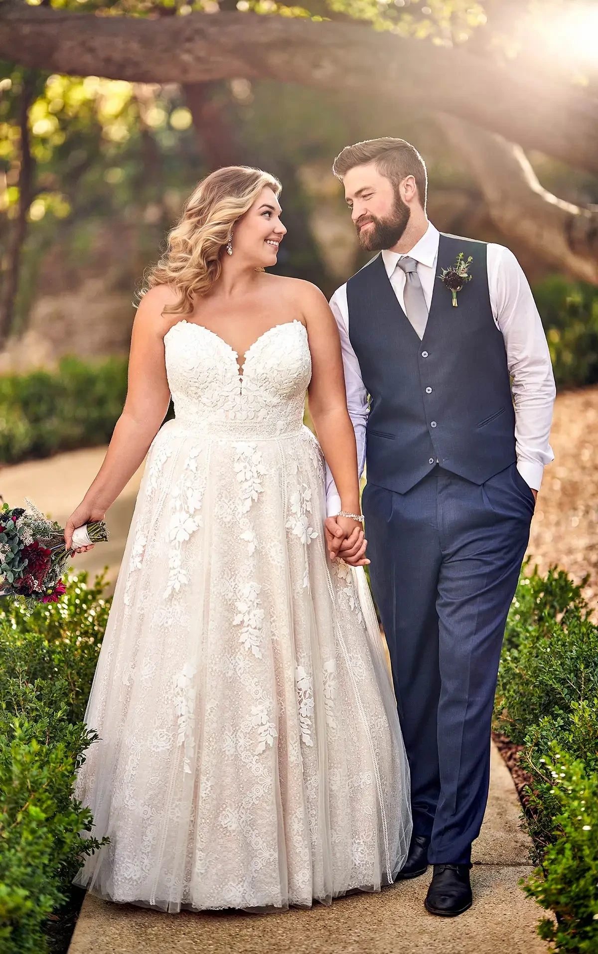 Bride wearing a plus-size strapless lace wedding gown with a sweetheart neckline and full A-line skirt, walking hand in hand with the groom outdoors.