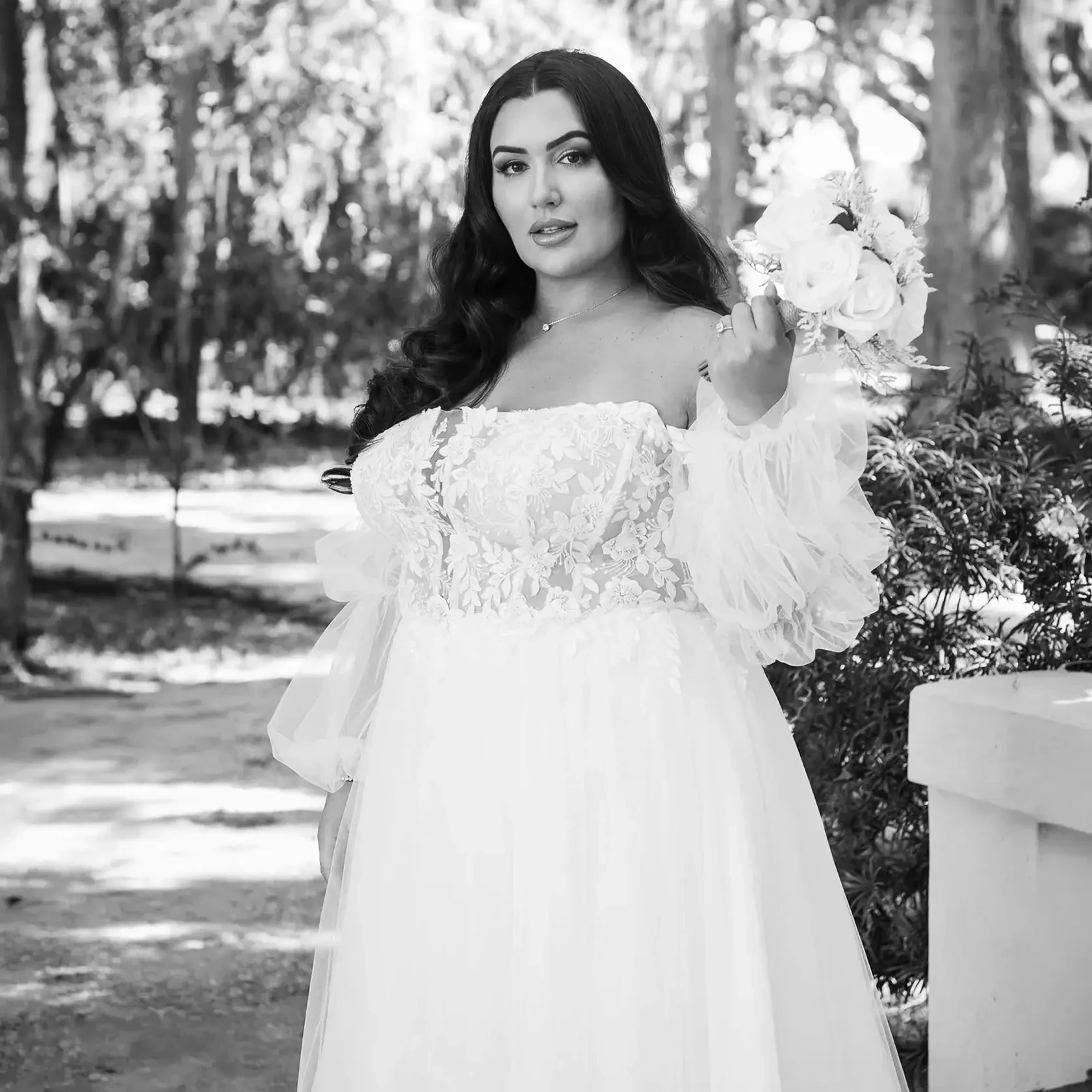Plus-size bride wearing a lace wedding gown with sheer puff sleeves while holding a white bridal bouquet outdoors.