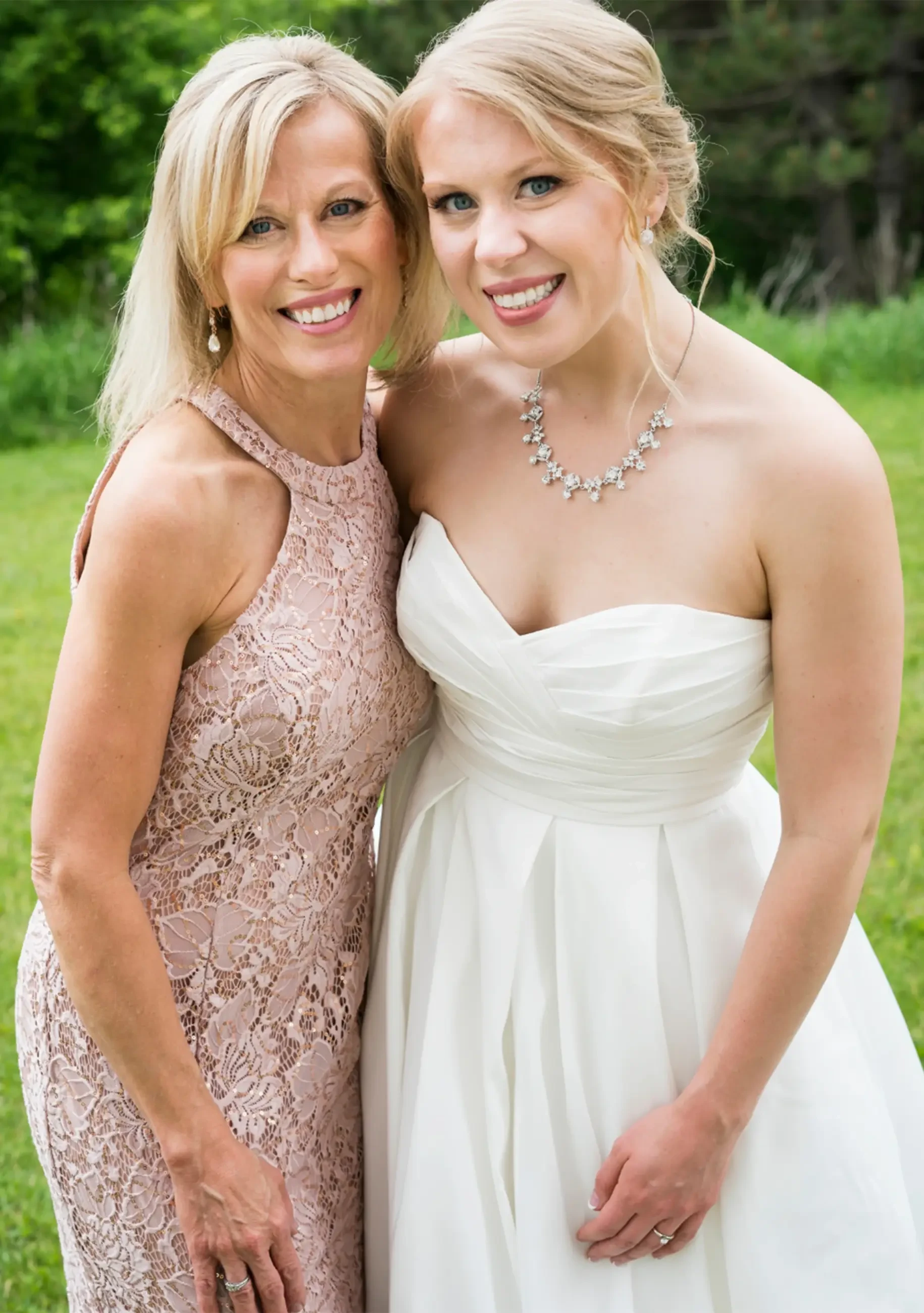 Bride in a strapless white wedding gown posing outdoors with her mom wearing a blush mother of the occasion lace dress.