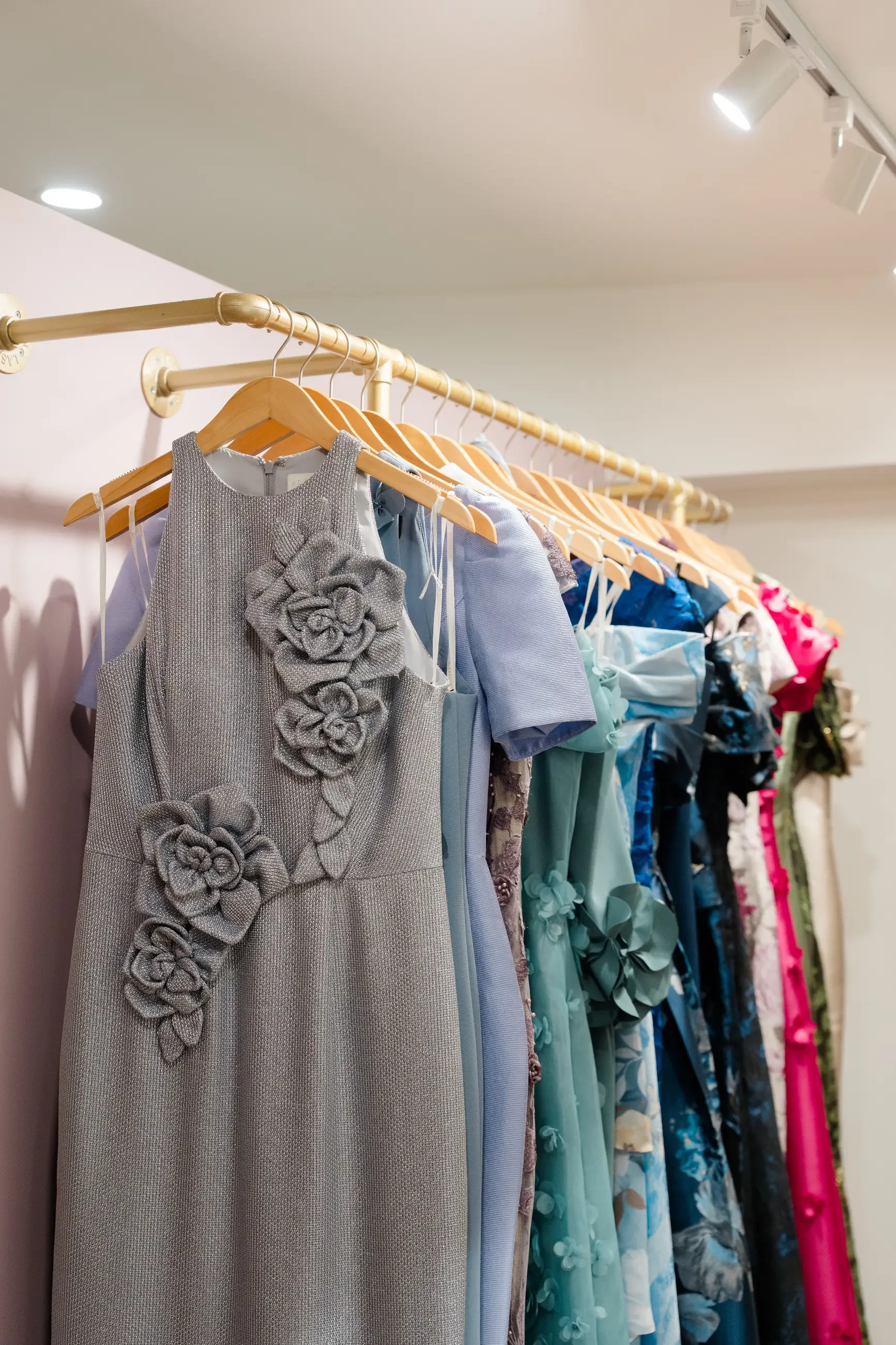 A rack of elegant occasion dresses in soft blue, silver, floral, and vibrant tones displayed inside a boutique.
